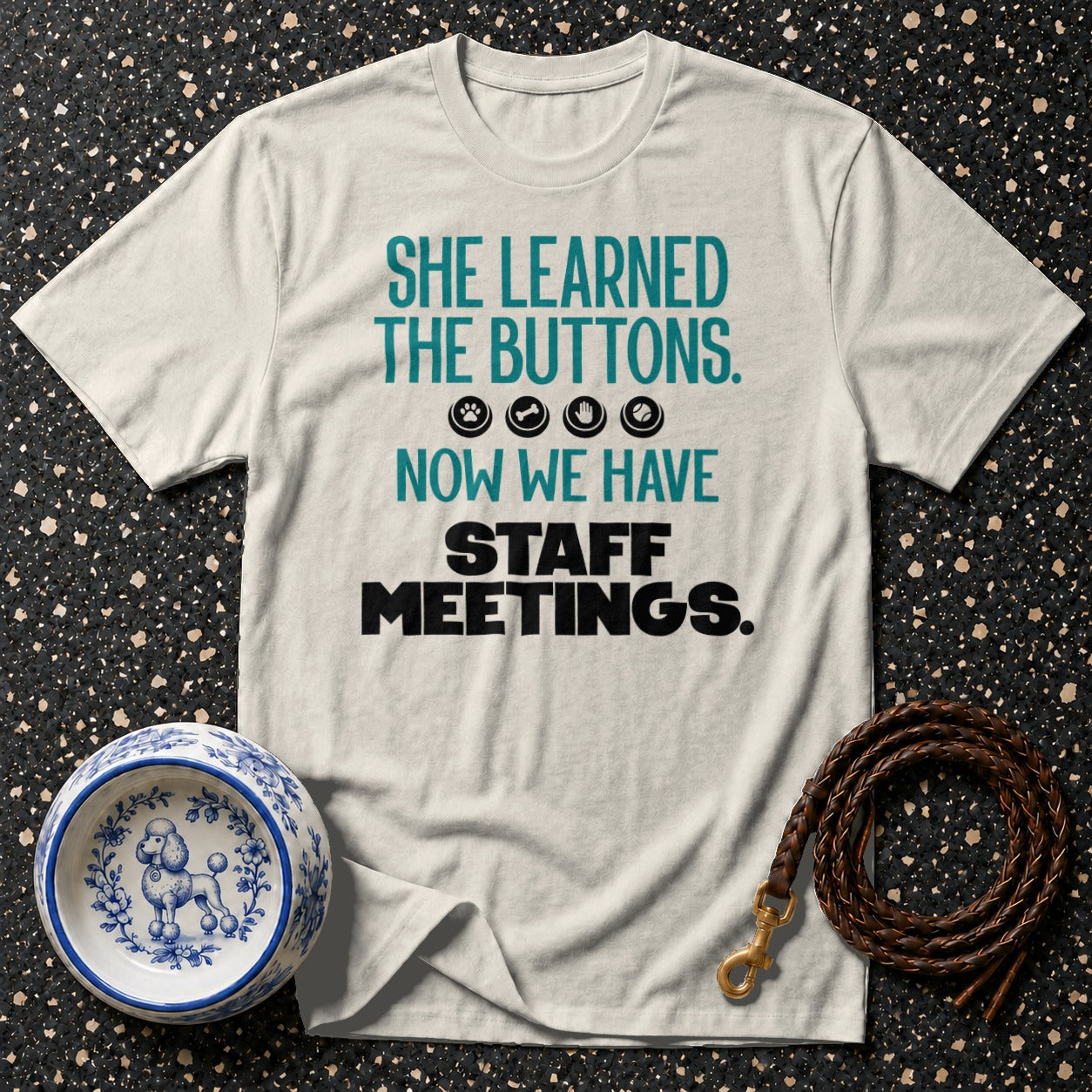 Light cream t-shirt with the text "She learned the buttons. Now we have staff meetings."