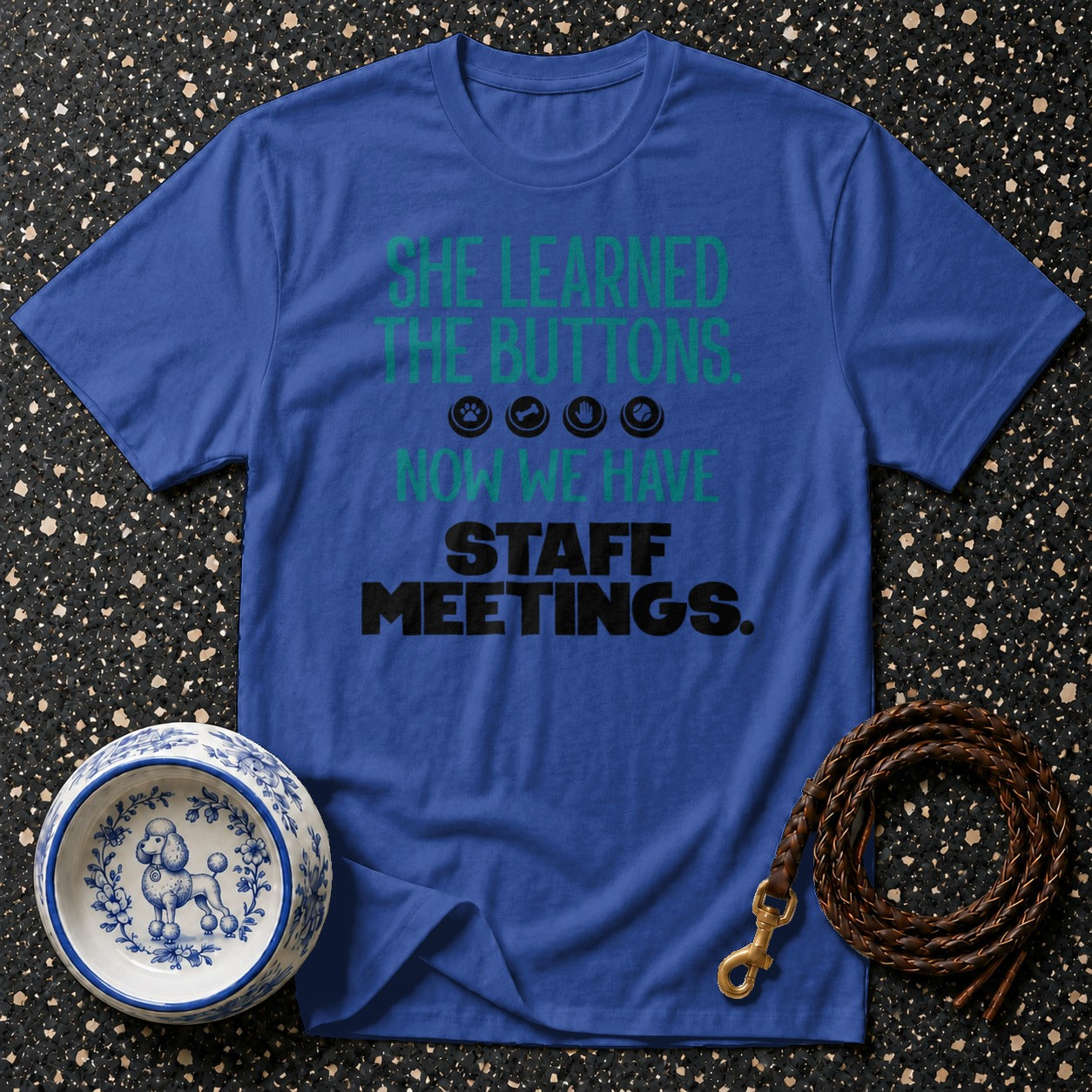 Blue t-shirt with green text "SHE LEARNED THE BUTTONS." and black text "STAFF MEETINGS.".