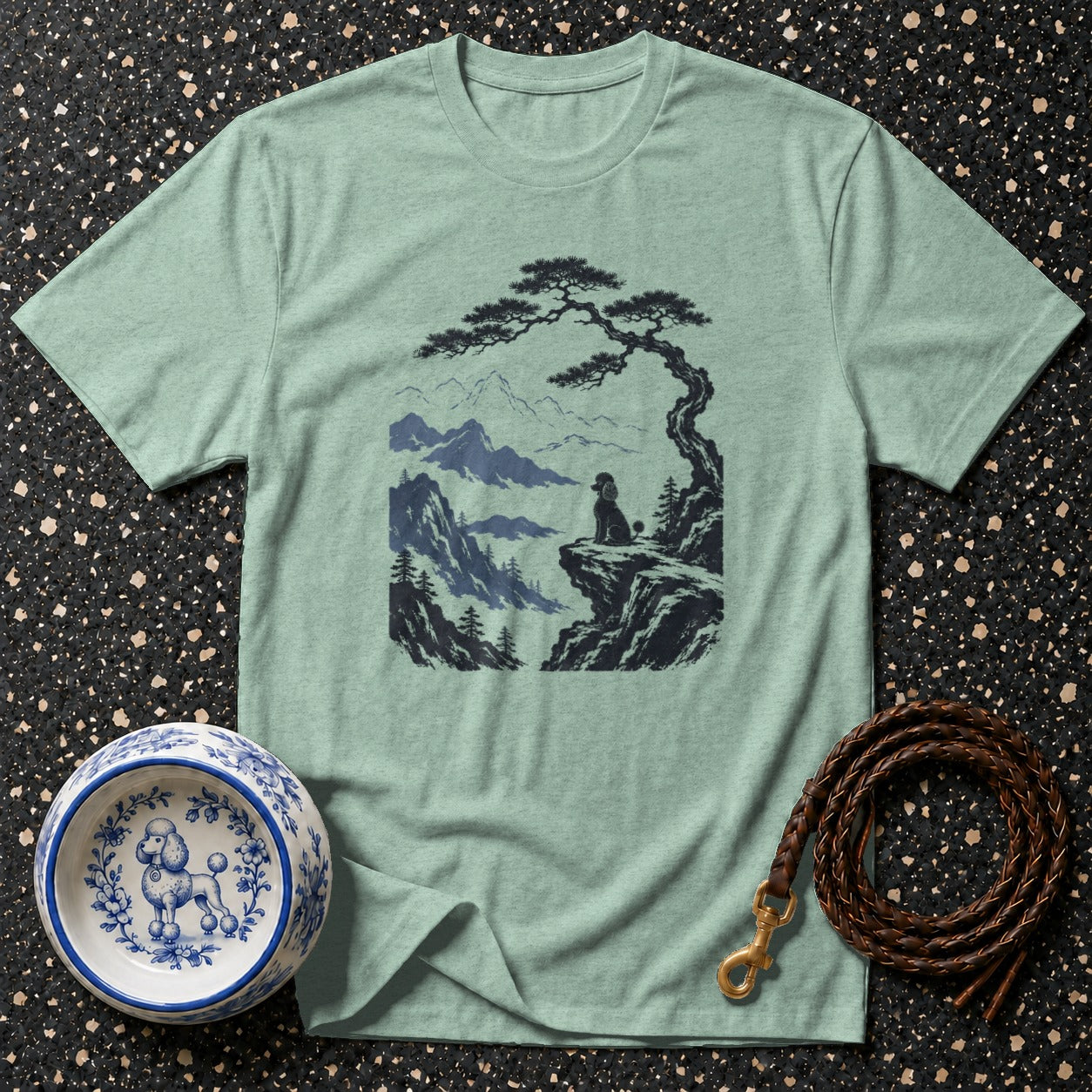 Light green t-shirt with graphic of poodle on cliff under pine tree and mountains.