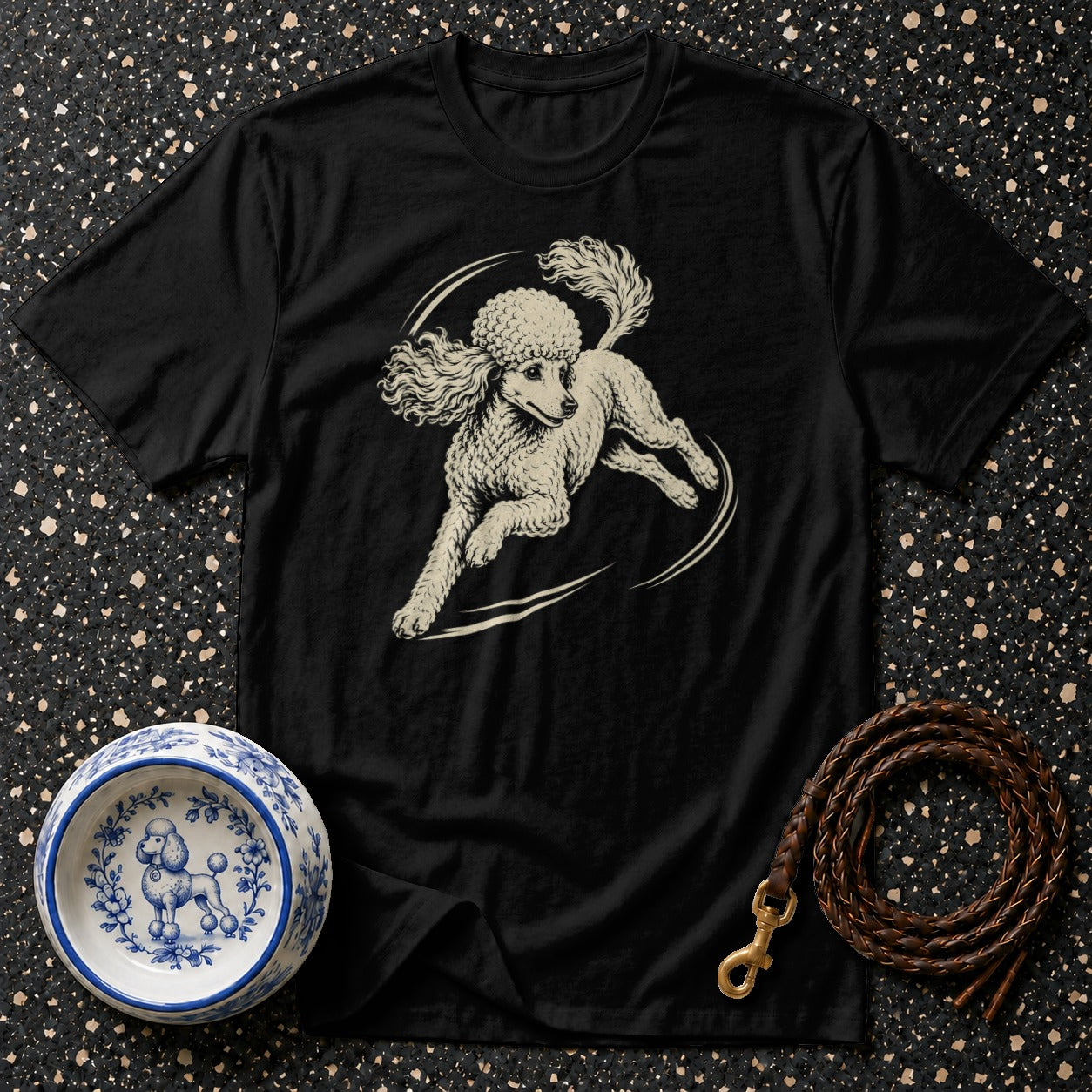 Black t-shirt with a cream-colored poodle illustration, beside a poodle-decorated bowl and leather leash.