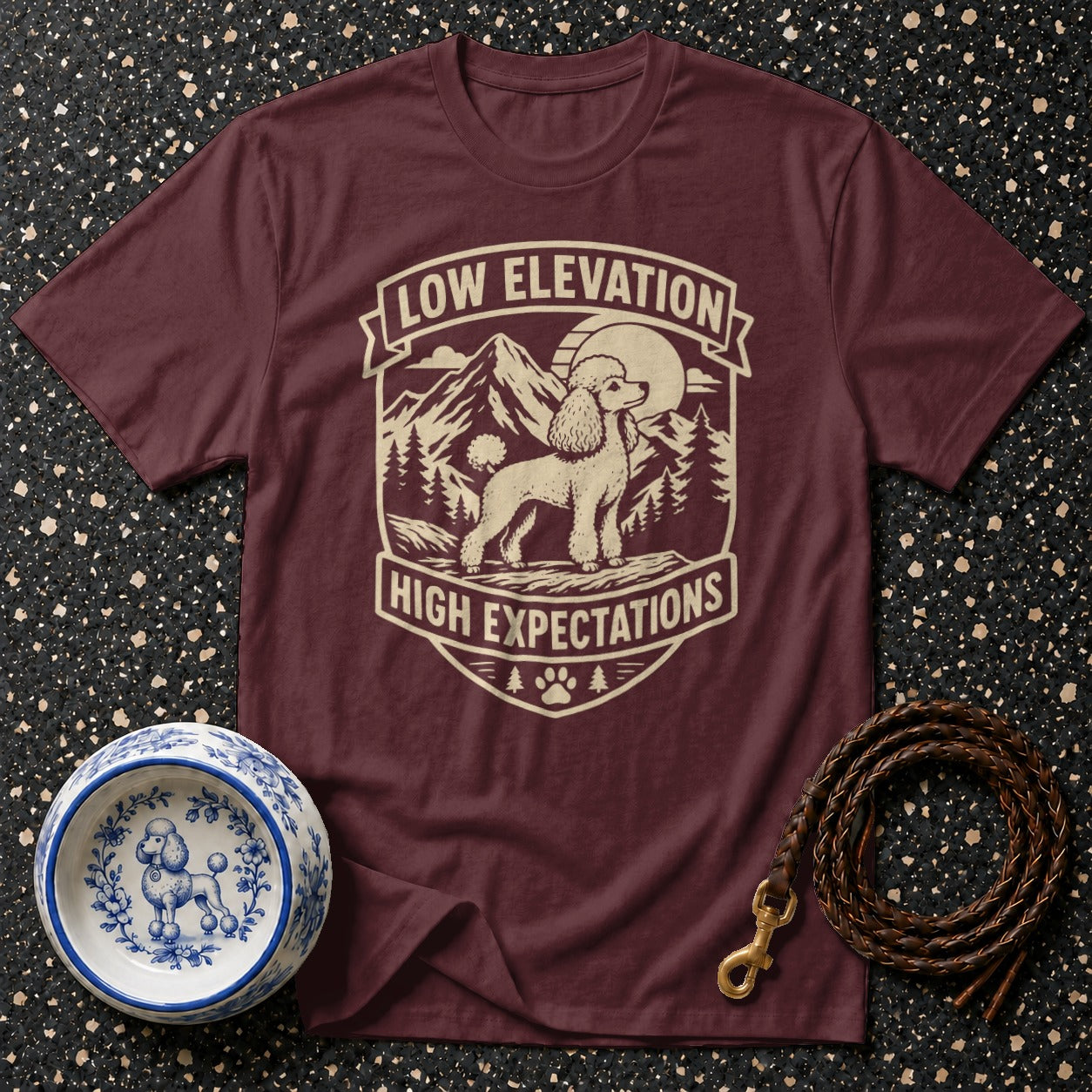 Burgundy t-shirt with a cream graphic of a poodle in a mountain scene, text reads "LOW ELEVATION HIGH EXPECTATIONS."