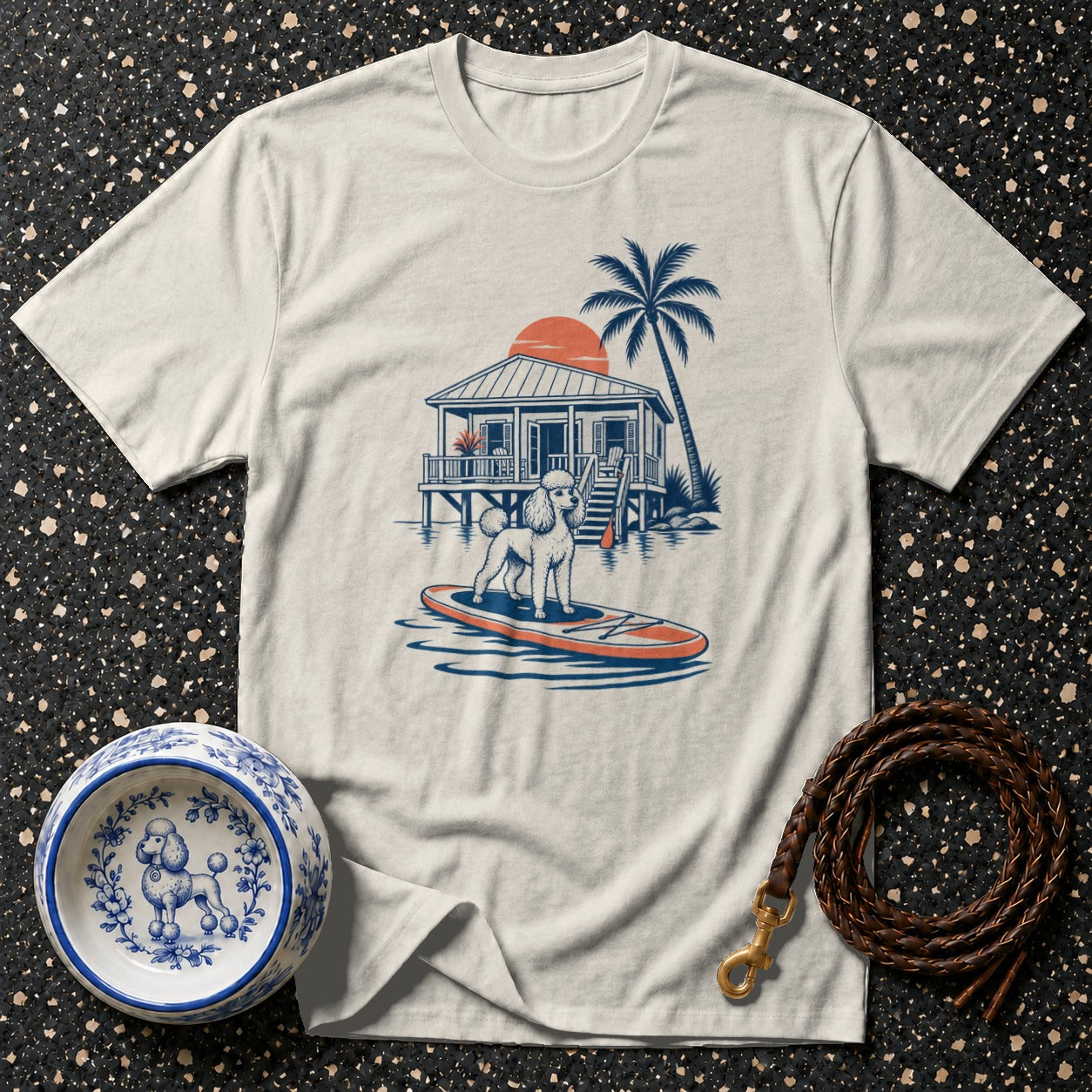 T-shirt graphic of a poodle on a paddleboard in front of a stilt house.