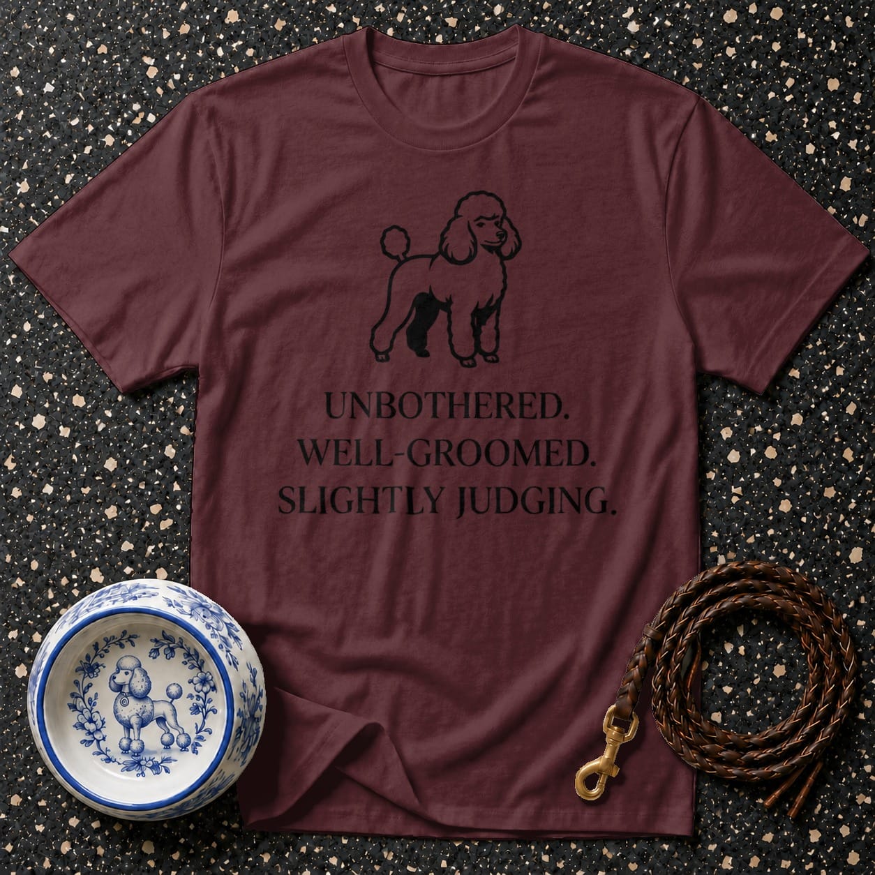 Maroon t-shirt with black poodle graphic and text reading "UNBOTHERED. WELL-GROOMED. SLIGHTLY JUDGING."