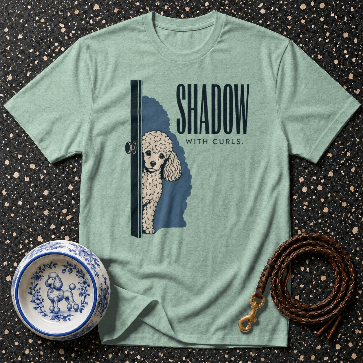 Light green t-shirt with graphic of poodle peeking around a door, text "SHADOW WITH CURLS."