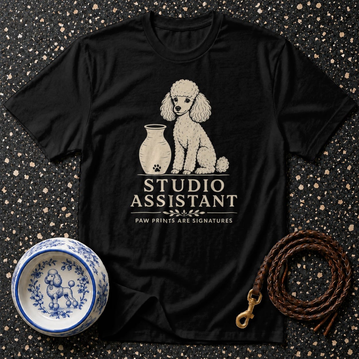 Black t-shirt with white poodle graphic, vase, and text "STUDIO ASSISTANT".