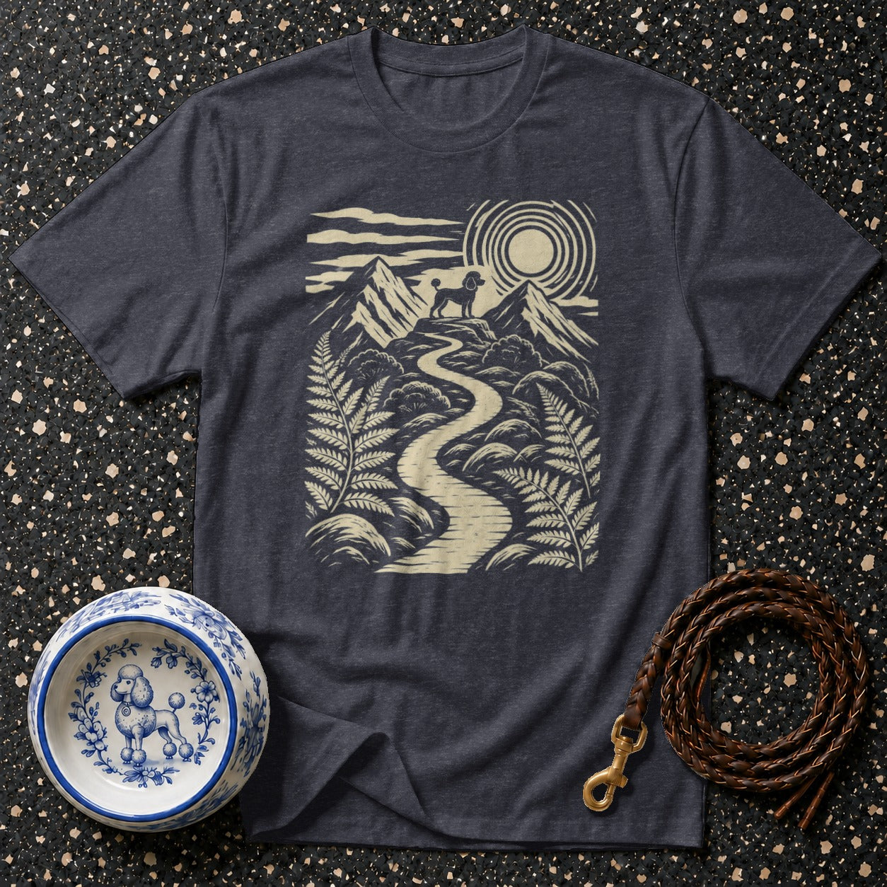 Dark t-shirt with a poodle on a mountain path design, next to a decorative dog bowl and leash.
