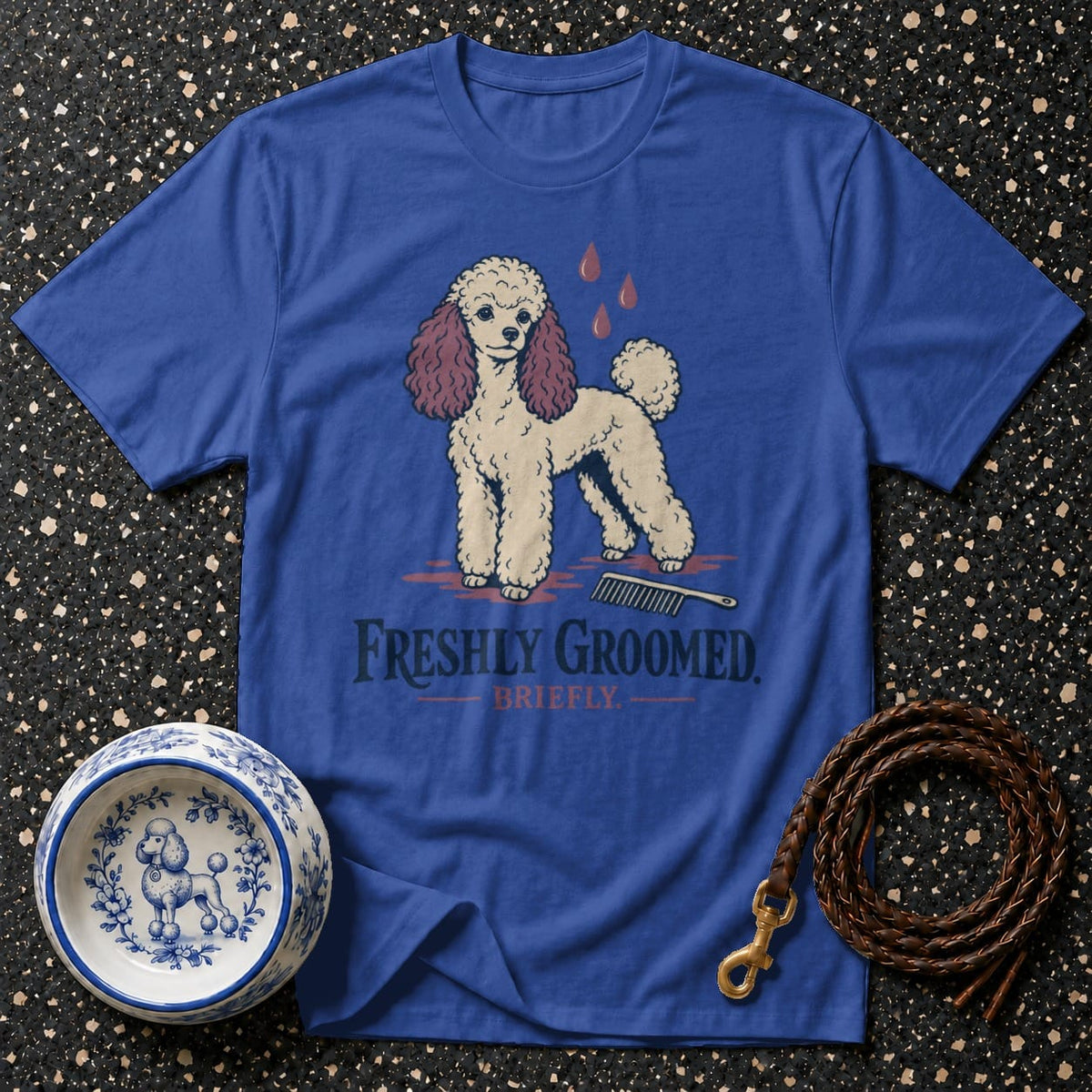 Blue t-shirt with illustration of a poodle, comb, and blood drops. Text reads "FRESHLY GROOMED. BRIEFLY."