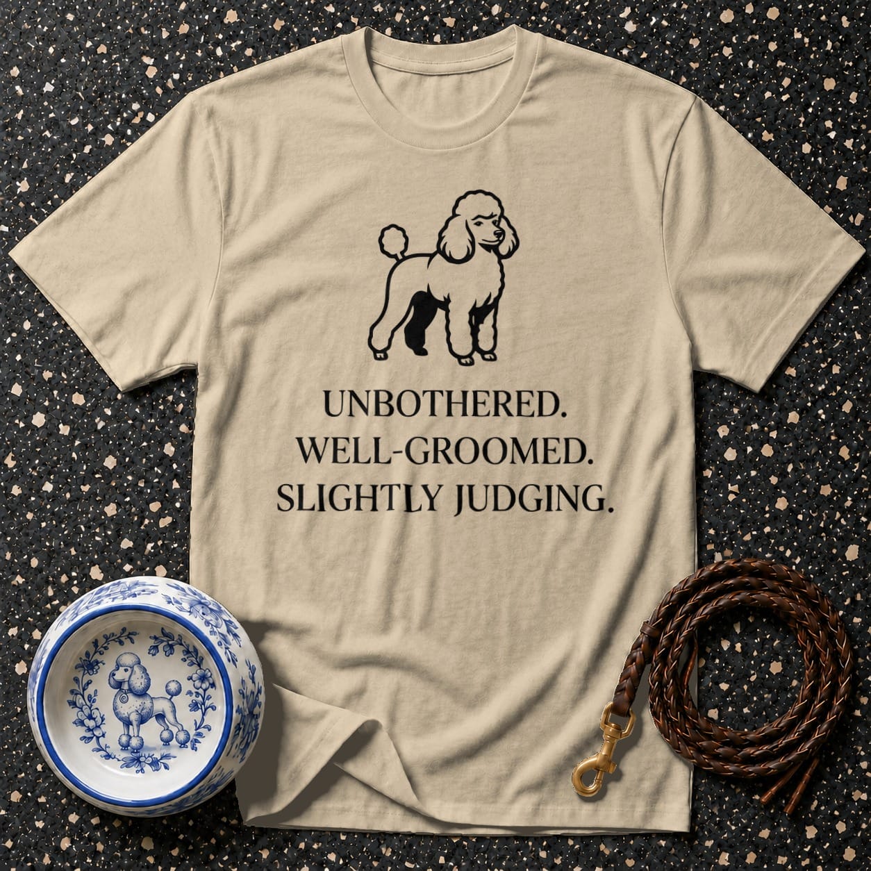 Light tan t-shirt with a poodle graphic and "UNBOTHERED. WELL-GROOMED. SLIGHTLY JUDGING." text.