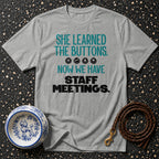 Grey t-shirt with text: "SHE LEARNED THE BUTTONS. ?�¾?¦´?Ž¾ NOW WE HAVE STAFF MEETINGS."