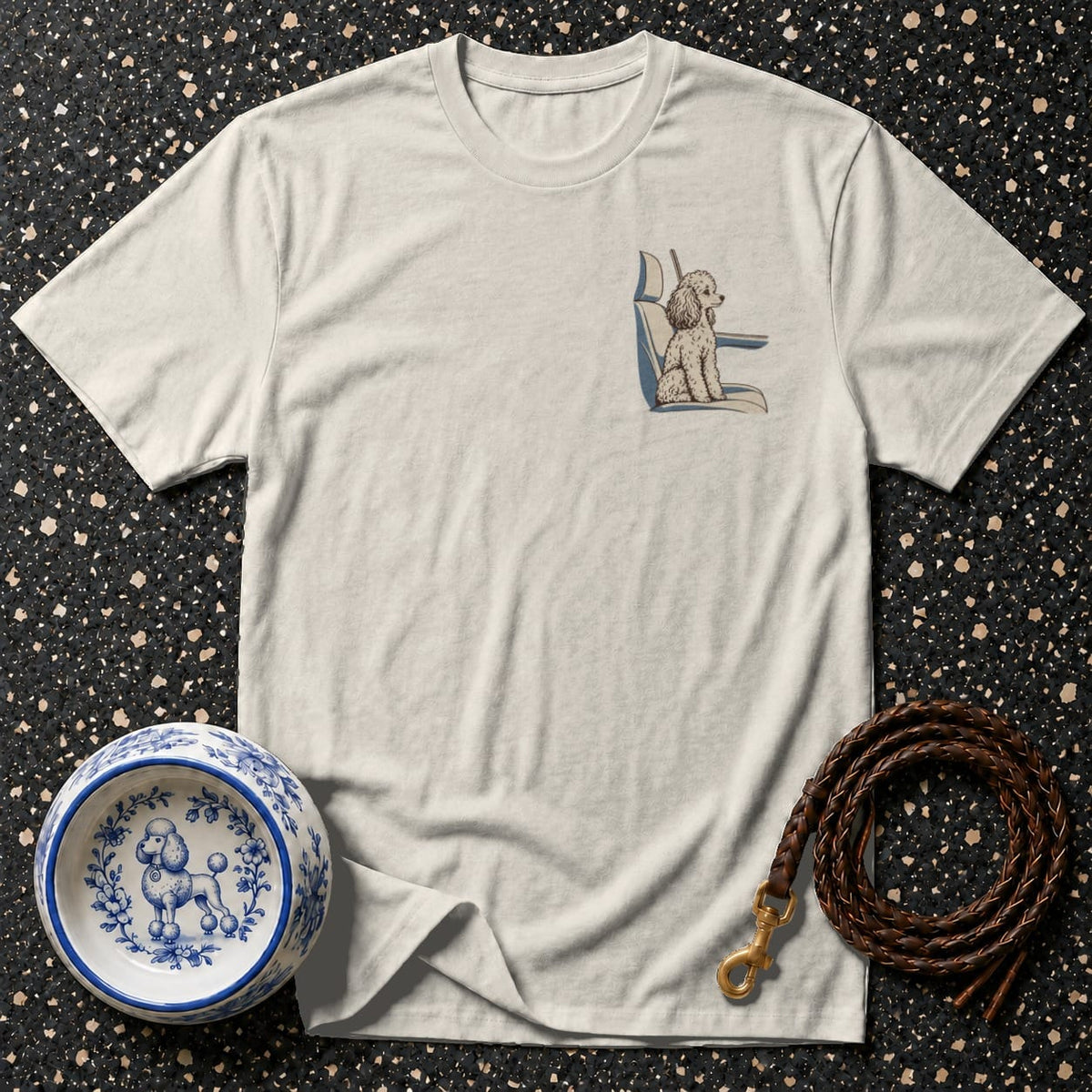 Off-white crewneck t-shirt featuring a small chest graphic of a poodle sitting in a car seat, laid flat on a speckled surface with a patterned pet bowl and a brown leather leash.