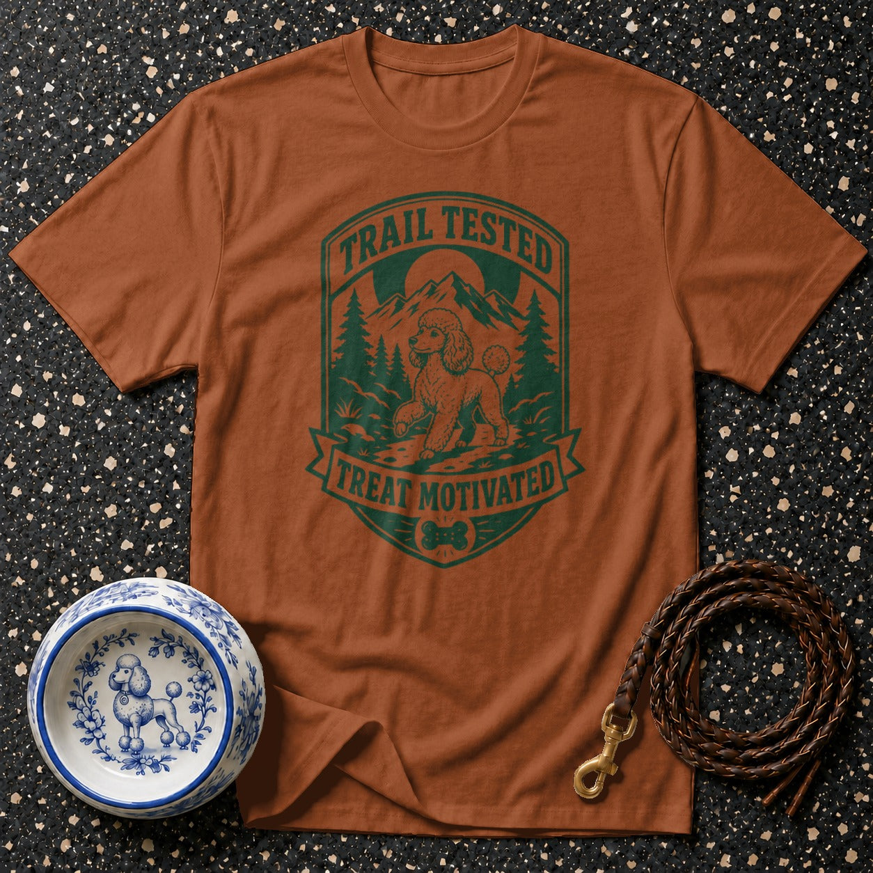Brown t-shirt with green graphic of a poodle on a trail, mountains, and trees.