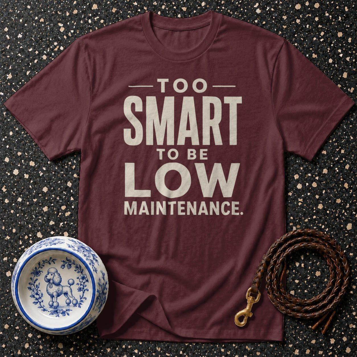Maroon t-shirt with "Too Smart To Be Low Maintenance" text, dog bowl, and leash on speckled background.
